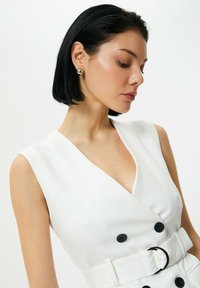 Vestido blanco con escote en V profundo, diseño sin mangas, botones negros y un cinturón como acento. Tela suave con ligero brillo.