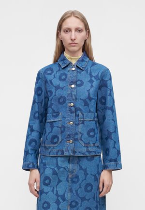 Femme portant une veste en jean bleue et une jupe avec un grand imprimé floral foncé, se tenant devant un fond clair uni.