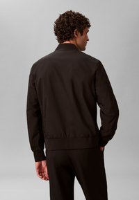 Veste bomber noire avec col et poignets côtelés, finition lisse et ceinture élastique, vue de dos.