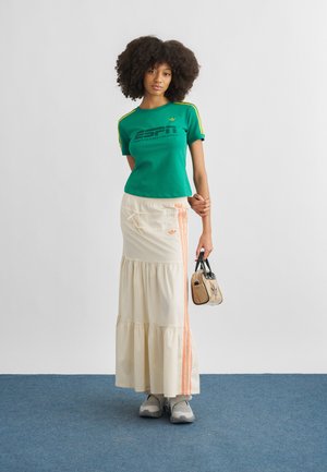 Vrouw met krullend haar, een groen ESPN-T-shirt, een crème lange rok met oranje strepen, grijze sneakers en een kleine beige handtas in haar hand.