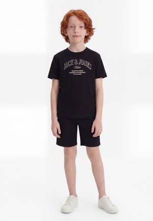 Garçon roux debout, portant un t-shirt noir Jack & Jones, un short noir et des baskets blanches sur un fond blanc uni.
