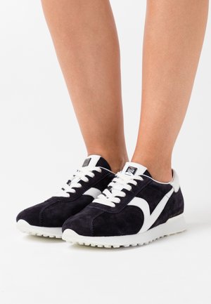 Navy suede sneakers with white accents, lace-up design, and a textured sole, featuring a logo patch on the tongue. Lightweight construction.