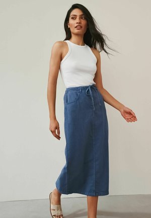 Frau mit langen dunklen Haaren, die ein weißes ärmelloses Oberteil, einen blauen Midirock mit Taschen und Taillenband sowie beigefarbene Sandalen trägt und drinnen geht.