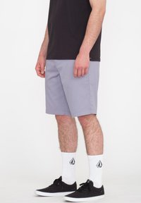 Grå shorts med en afslappet pasform, glat stof og ensfarvet. Båret med sorte sneakers og hvide sokker med logo, stående på en neutral baggrund.