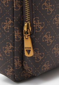 Sac à main marron à motif avec quincaillerie dorée, doté d'une fermeture éclair et d'un logo "GUESS" bien en vue sur la tirette de la fermeture. Matériau en cuir texturé.