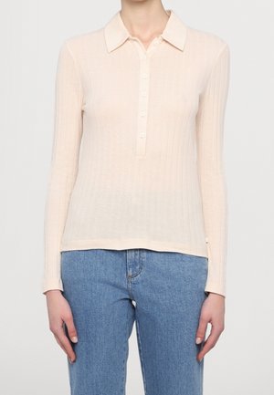 Beige ribbet langærmet polo-shirt med knaplukning, båret med blå denimjeans, centreret på overkrop og arme mod ensfarvet baggrund.