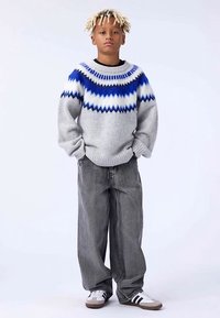 Γκρι πλεκτό πουλόβερ με μπλε γεωμετρικό σχέδιο, συνδυασμένο με άνετο γκρι τζιν και λευκά αθλητικά παπούτσια με μαύρες ρίγες. Ομαλή υφή συνολικά.