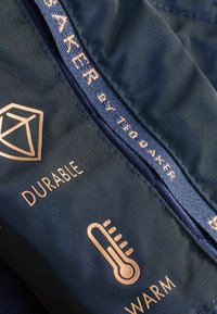 Tessuto navy con testo e icone dorate che indicano durata e calore. Presenta una zip texturizzata e il logo "TED BAKER by TED BAKER."