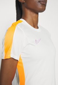 Valge sportlik t-särk lühikeste varrukatega, millel on oranžid mesh-küüred ja väike roosa Nike logo rinnal. Sile tekstuur.