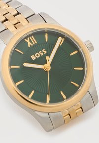 Χρυσοί και ασημένιοι μεταλλικοί ρολόι χειρός με πράσινο καντράν, χρυσούς δείκτες ώρας, λατινικό αριθμό XII και σήμα "BOSS", που δείχνει ώρα 9:03:02.