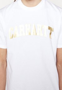 Valkoinen puuvillapaita, jossa on isot kultaiset tekstikirjaimet "CARHARTT" rohkealla, suurilla kirjaimilla rinnan kohdalla. Lyhyet hihat, pyöreä pääntie.