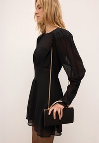 Svart klänning med genomskinliga långa ärmar, en åtsittande bodice och utsvängd kjol. Kombinerad med en liten svart clutch med en guldkedja.