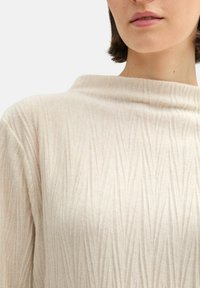 Haut beige clair avec une texture douce, présentant un col drapé et un motif à côtes verticales, mettant en valeur son design drapé.
