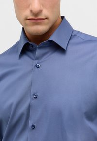 Zbliżenie mężczyzny w niebieskiej koszuli z button-up i spiczastym kołnierzykiem, na tle jasnego, jednolitego tła.