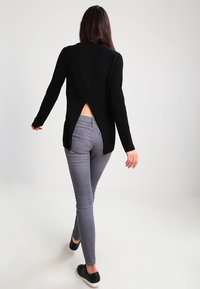 Mustan ribbipuseron, jossa on korkeampi takaosa, yhdistettynä slim fit -harmaisiin farkkuihin ja mustiin liukuvarpaisiin kenkiin.