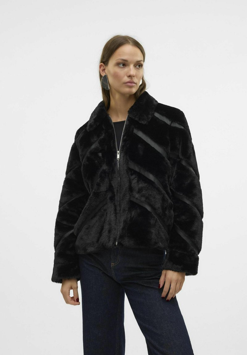 Czarna kurtka z sztucznego futra z zapięciem na zamek, z wyraźnym kołnierzem i skośnymi wzorami teksturowanymi. Nosi na sobie ciemną bluzkę i dżinsy.