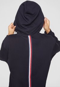 Personne ajuste la capuche d’un sweat sombre avec une bande verticale rouge, blanche et bleue dans le dos.