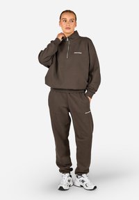 Brązowa bluza z wysokim kołnierzem i zamkiem, zestawiona z pasującymi spodniami dresowymi. Posiada białe akcenty logo oraz wygodne dopasowanie.
