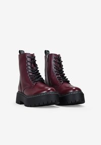 Botas de cuero burdeos de cordones con suelas gruesas negras, ojales plateados, cremalleras laterales y tiradores negros con letras blancas.