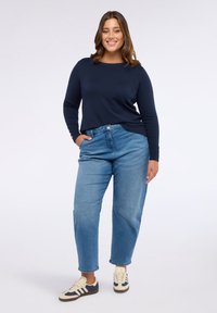 O femeie zâmbitoare stă cu mâna în buzunar, purtând un pulover bleumarin, blugi desermi și adidași albi cu dungi albastre, pe un fundal alb.