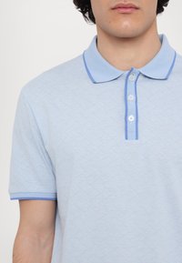 Polo bleu clair avec un motif en zigzag texturé, col et bord de manches contrastants en bleu plus foncé, avec trois boutons.
