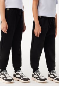 Schwarze Fleece-Sweatpants mit elastischem Bund, versehen mit einem kleinen grünen Lacoste-Logo. Kombiniert mit schwarz-weißen Sneakers.