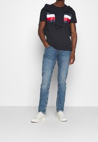 Camiseta azul marino con rayas horizontales rojas y blancas, combinada con vaqueros azul claro. Zapatillas blancas completan el conjunto. La textura parece suave.