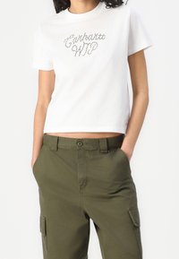 Valkoinen puuvillainen t-paita, jossa on lyhyet hihat ja lyhyempi malli. Edessä on tekstuurilla varustettu hopeinen helmi-logo, jossa lukee "Carhartt WIP".