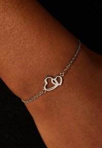 Zilveren hartvormige armband met een dunne ketting, rustend op een pols. Het hartontwerp heeft een open lus, die contrasteert met de huidskleur.