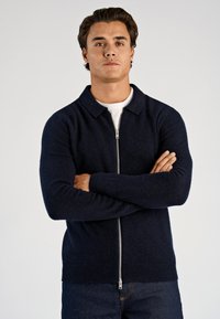 Granatowy sweter na zamek wykonany z fakturowanej dzianiny, z kołnierzem, długimi rękawami oraz ściągaczami na mankietach i dole. Noszony na białej koszuli.