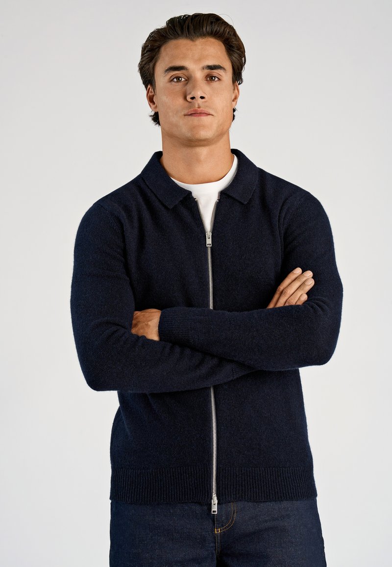 Granatowy sweter na zamek wykonany z fakturowanej dzianiny, z kołnierzem, długimi rękawami oraz ściągaczami na mankietach i dole. Noszony na białej koszuli.