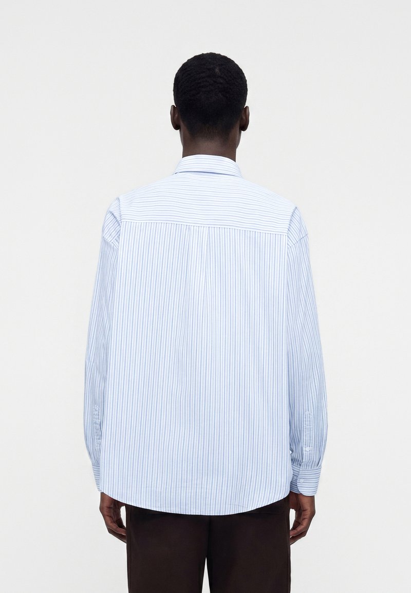 Uomo che indossa una camicia a maniche lunghe rigata verticalmente azzurro chiaro e bianco, in piedi di spalle su uno sfondo bianco semplice.