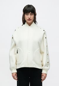 Νεαρός με σκούρα μαλλιά, που φοράει λευκό μπουφάν με κυκλικές εγκοπές στα μανίκια και μαύρο παντελόνι, κοιτάζει μπροστά με φόντο ένα απλό φόντο.