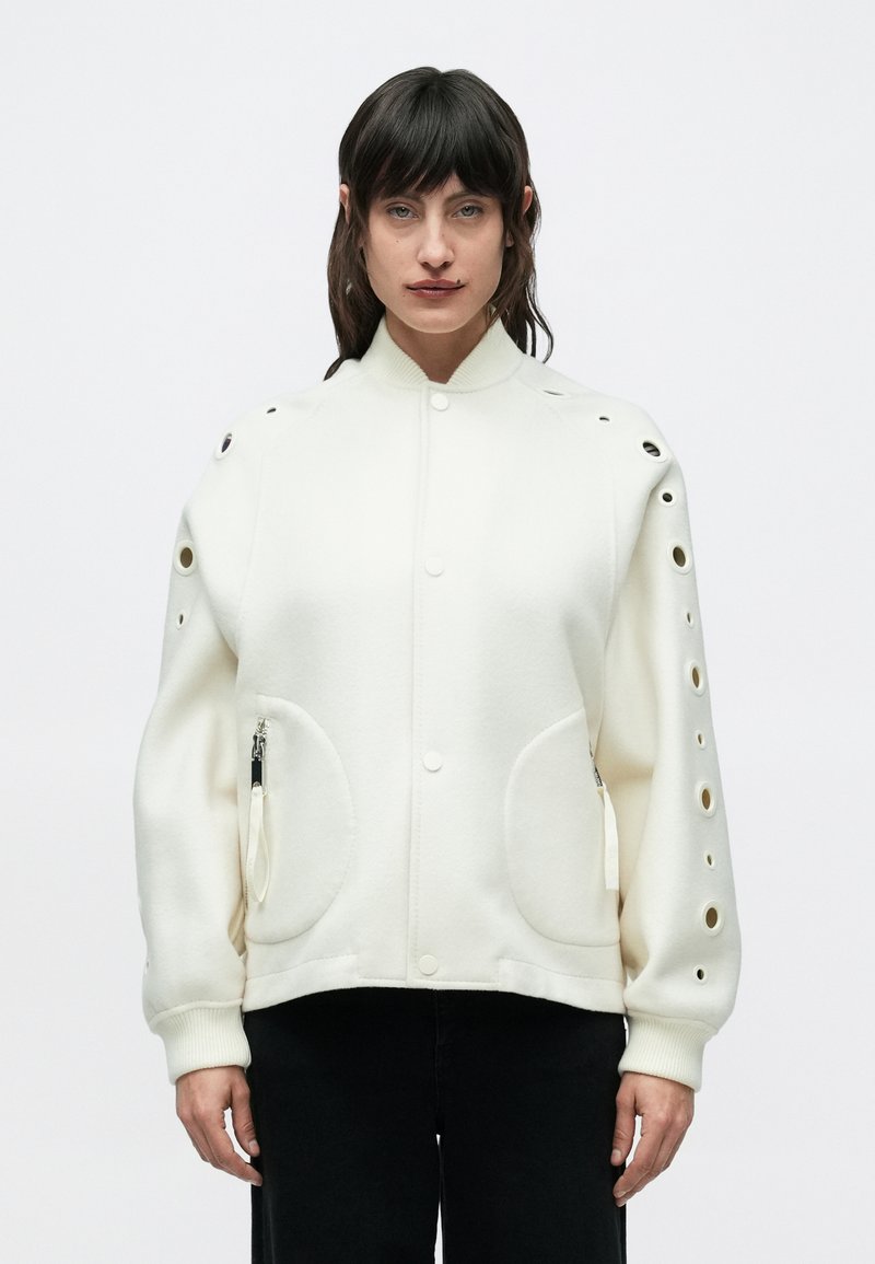 Νεαρός με σκούρα μαλλιά, που φοράει λευκό μπουφάν με κυκλικές εγκοπές στα μανίκια και μαύρο παντελόνι, κοιτάζει μπροστά με φόντο ένα απλό φόντο.