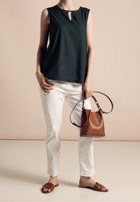 Ciemnozielony bezrękawnik z dekoltem w kształcie łezki, zestawiony z białymi spodniami w paski, brązową torebką i brązowymi sandałami na płaskim obcasie.