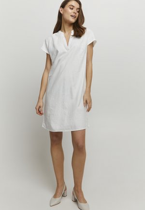 Robe en lin blanc avec une coupe décontractée, des manches courtes et un décolleté en V. Le tissu a une texture subtile et arrive au milieu de la cuisse. Portée avec des talons beiges.