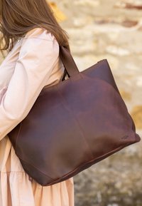 Brown leather tote bag with a smooth texture, rounded shape, and stitching detail. It features dual handles for easy carrying.