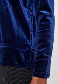 Top in velluto blu navy con una superficie liscia e lucida. Presenta un design aderente e maniche lunghe, abbinato a una gonna nera plissettata.