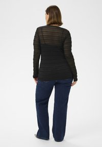 Femme aux cheveux bruns courts portant un haut noir transparent à manches longues et un jean bleu foncé, se tenant de dos sur un fond blanc.