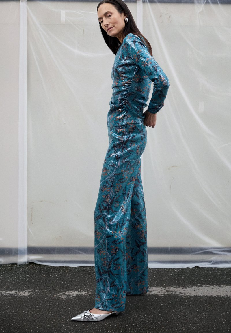 Ολόσωμη φόρμα από τυρκουάζ γκλίτερ με μακριά μαν ίκια και ρυτιδωμένη λεπτομέρεια στο πλάι, διακοσμημένη με φυτικά μοτίβα σε χαλκοειδείς αποχρώσεις. Συνδυασμένη με ασημί παπούτσια με μυτερή μύτη.