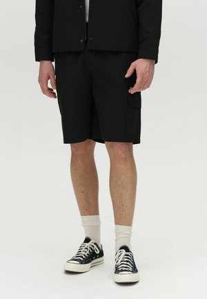 Pantalones cortos de carga negros con bolsillos laterales, hechos de una tela ligera. Llevados con zapatillas negras y calcetines blancos hasta la mitad de la pierna. Diseño y ajuste simples.