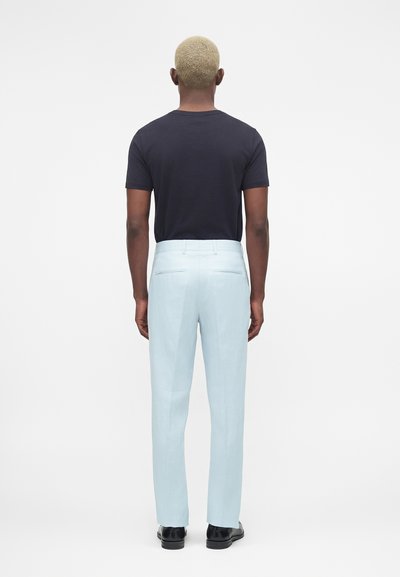 Άνδρας με κοντά ξανθά μαλλιά που φοράει μαύρο μπλουζάκι, ανοιχτό μπλε παντελόνι και μαύρα παπούτσια, στέκεται με την πλάτη προς τη λευκή επιφάνεια.