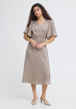 Kobieta z długimi kręconymi włosami w taupe satynowej sukience midi z rękawami motylkowymi i beżowymi szpiczastymi szpilkami, stojąca na białym tle.