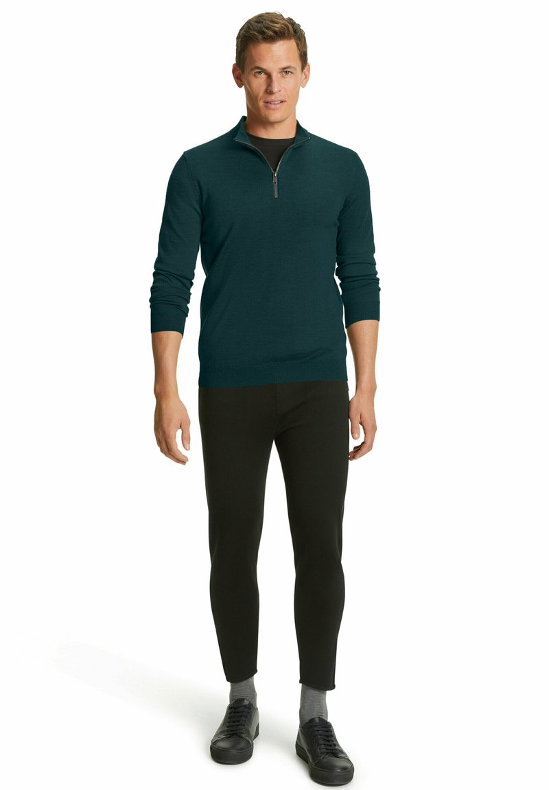 Maglione verde azzurro con collo a zip e maniche lunghe, abbinato a pantaloni neri attillati e sneakers nere. Il tessuto appare liscio e strutturato.