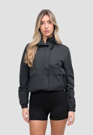 Kobieta o długich blond włosach, ubrana w czarną kurtkę z kapturem i czarne szorty, stojąca na tle jednolitego szarego tła.