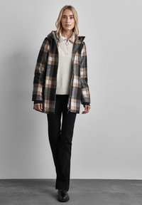 Giacca a quadri nera, beige e grigia con cappuccio, indossata su un maglione crema. Abbinata a pantaloni neri e scarpe scure. Tessuto morbido.