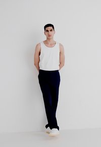 White tank top, dark navy trousers, and white sneakers. Model stands against a plain background, arms relaxed at sides. Smooth fabric, tailored fit.