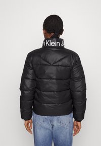 Chaqueta acolchada negra con un diseño geométrico acolchado, que cuenta con un cuello alto con un logo blanco. Llevada sobre unos vaqueros azules de mezclilla.