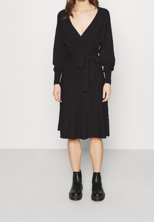 Žena v černých zavinovacích šatech s výstřihem do V, dlouhými rukávy a páskem v pase, v kombinaci s černými kotníkovými botami na neutrálním pozadí.