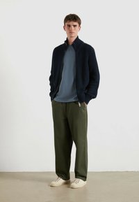 Maglione zip-up a coste blu navy sopra una t-shirt azzurro chiaro, abbinato a pantaloni larghi verde oliva e sneakers beige, posizionato su uno sfondo semplice.